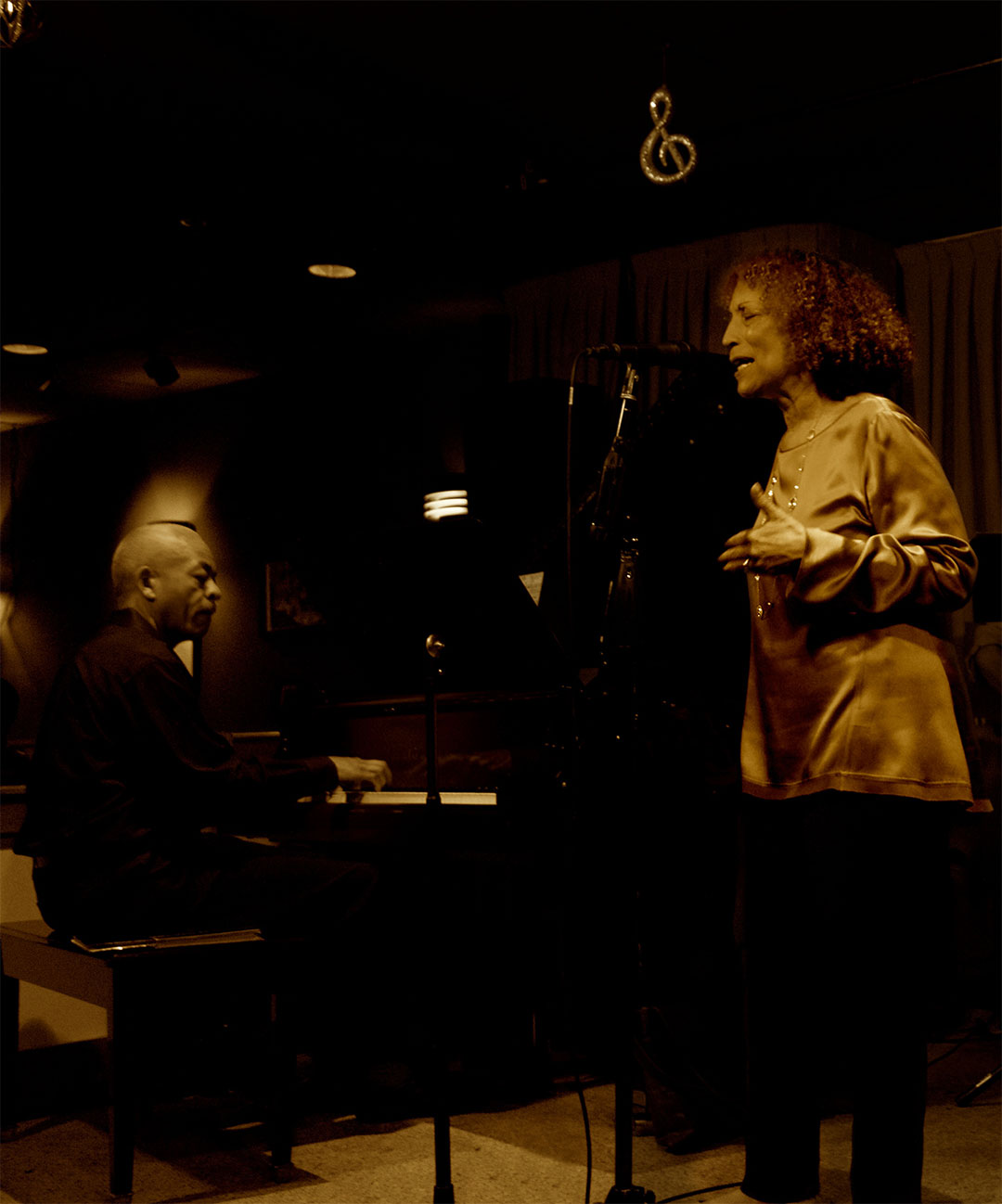 Adegoke Steve Colson at the piano and Iqua Colson singing at a microphone in a dimly lit club, photographed in warm sepia tones.